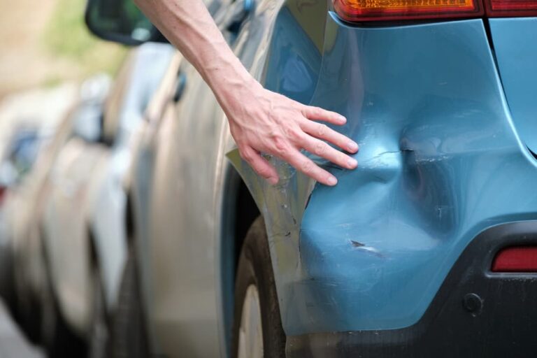 A driver examines the dented fender of a car parked on the side of a city street. This image highlights road safety and the importance of vehicle insurance.