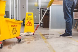 A "Caution Wet Floor" sign warns passersby as a string mop cleans a wet concrete floor.