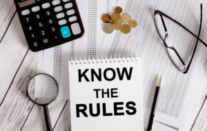 White notepad with 'KNOW THE RULES' written on it, placed next to a calculator, cash, glasses, magnifying glass, and pen. Business concept.