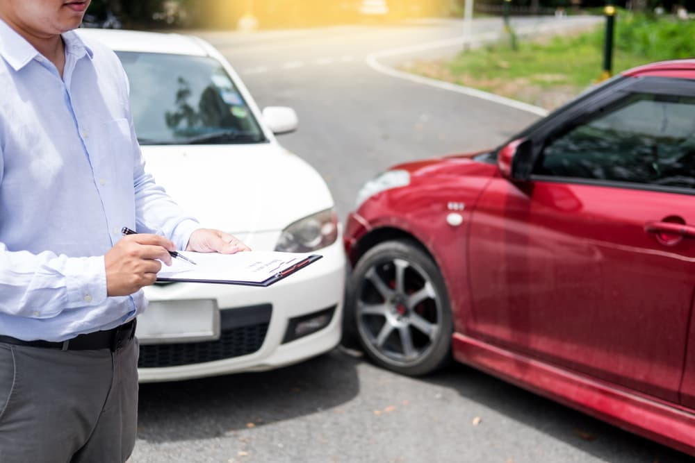 An insurance agent is writing on a clipboard while inspecting a rental car that is being assessed and processed for an accident claim.