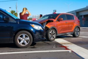 Two cars crashed at an intersection, with severe damage visible. An upset male driver inspects the wreckage, while a 'wrong way' sign is seen in the background.