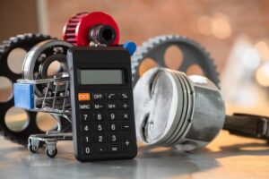 Car repair costs concept: Calculator, engine piston, and bearings in a shopping cart on a metal workbench background, close-up.